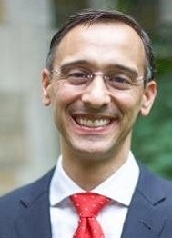 Alan Kahn, a man with gray and dark hair wearing a blue suit and red tie wearing glasses.