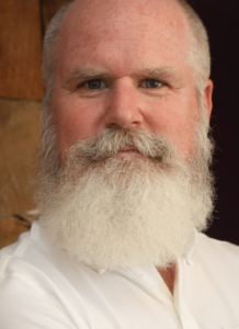 Justin Rood, an older man with white hair and beard wearing a white shirt.