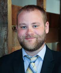 Tim Stretton, a dark haired man with a beard wearing a light blue shirt and yellow, blue, and white tie.