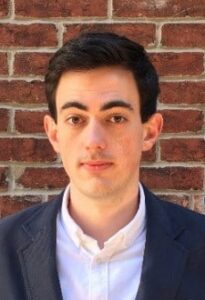 Elliot Smith, a man with short dark hair standing in front of a brick background wearing a dark sport jacket and white shirt.
