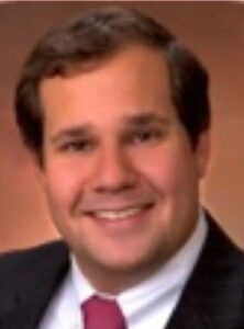 John Strom, a dark haired man wearing a dark suit and red tie.