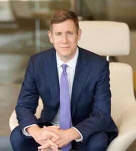 Jonathan Skladany, a dark haired man sitting on a tan seat wearing a dark blue suit and light blue tie.